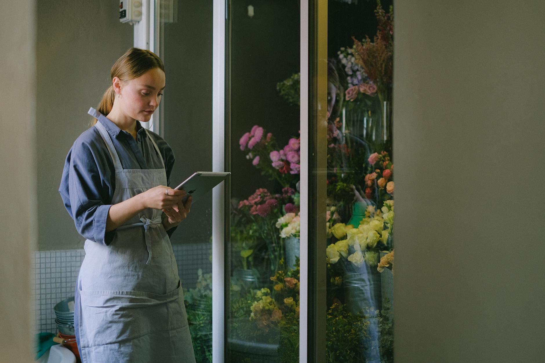 Kwiaciarka w fartuchu korzysta z tabletu obok stoiska z kwiatami